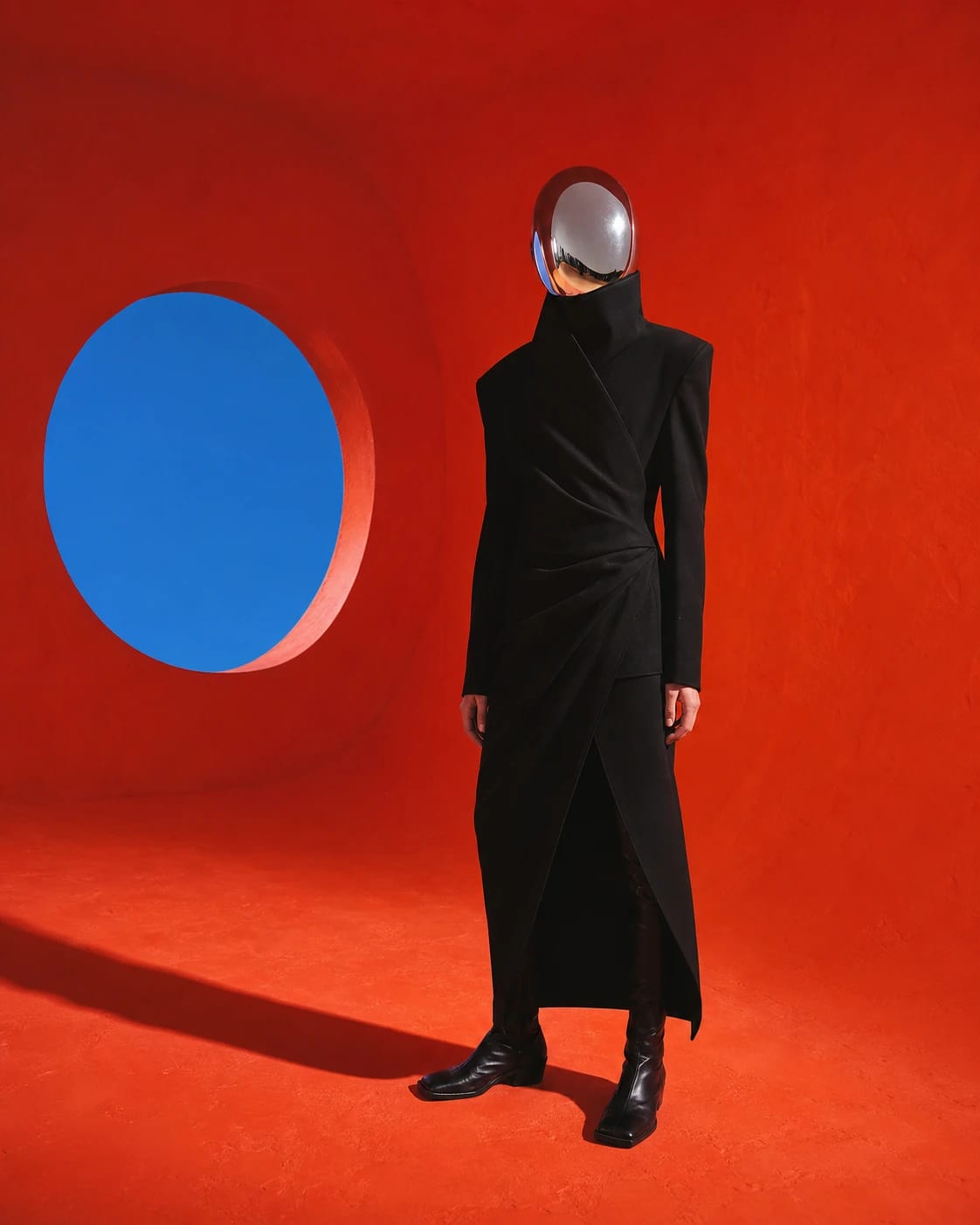 A figure in an architectural black garment and chrome visor standing in a vermilion room.
