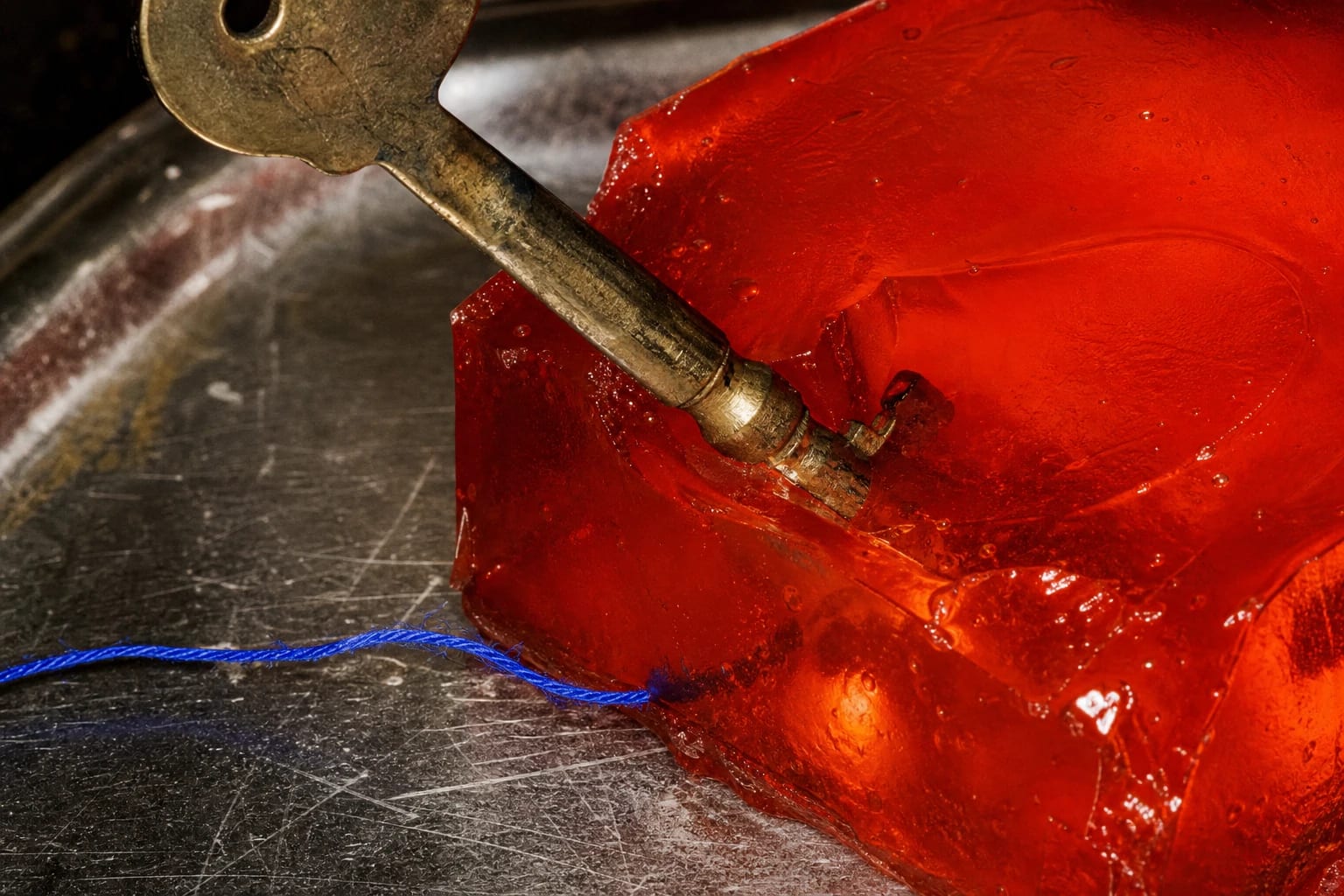 An old brass key pressed into translucent vermilion gelatin on a scratched metal surface.