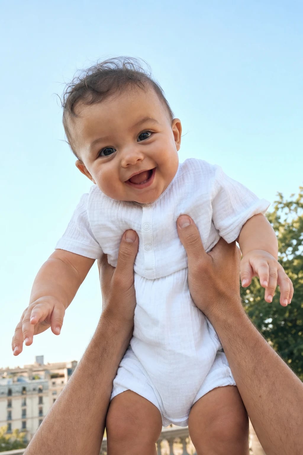 A smiling baby in white held aloft beneath an open blue sky.